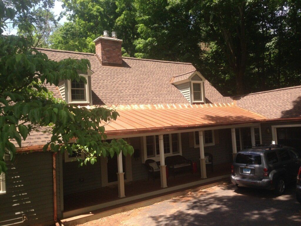 Brown-roofed house with copper-colored porch roof; car parked on right; trees in background.