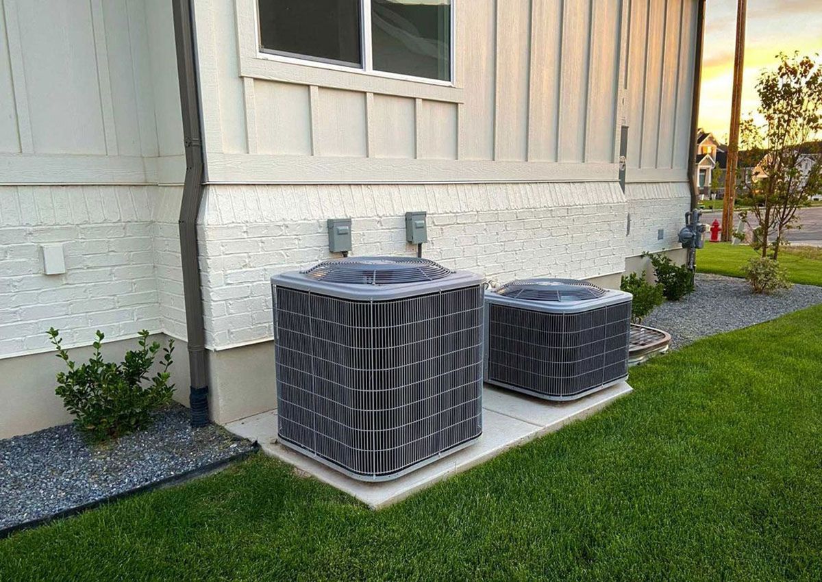 Two outdoor air conditioning units beside a house on a small concrete pad beside green lawn.