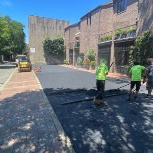 Workers paving a brick-lined street beside buildings, with a small road roller on the left.