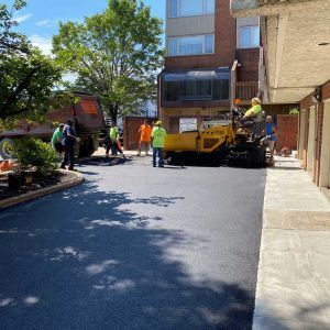 Construction crew paving a courtyard beside a brick building, with a yellow road paver and workers in safety vests.