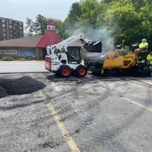 Road crew using a skid steer to load hot asphalt into a paver on a smoky street repair site