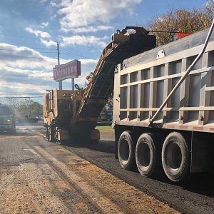 Road milling machine loading asphalt into a dump truck on a sunny roadside.