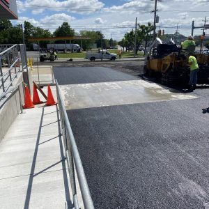 Road crew paving a driveway ramp beside a concrete sidewalk, with cones and workers using asphalt equipment.