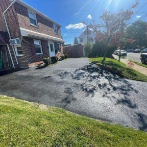 Suburban driveway beside brick house, green lawn, tree shadows, and bright sunny sky