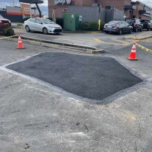Fresh black asphalt patch in a parking lot, bordered by orange cones near parked cars and a building.