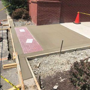 Fresh concrete sidewalk ramp with pink tactile warning strip beside a brick wall and orange cone