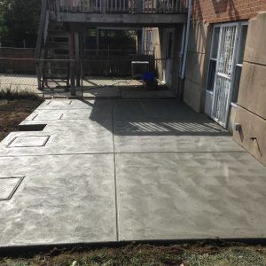 Concrete backyard patio beside a building, with steps, a gate, and shadows from the railing.