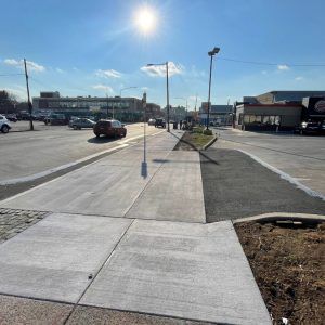 Sunny sidewalk beside a parking lot and storefronts, with a streetlight and clear blue sky.
