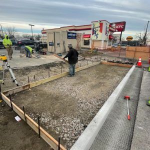 Workers building a concrete walkway beside a KFC, with gravel, forms, and construction cones.