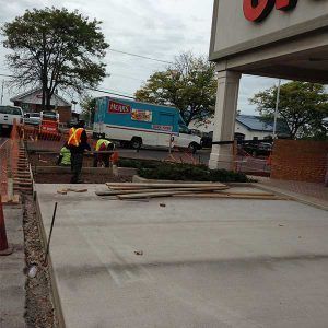 Workers repairing a concrete walkway outside a store, with a delivery truck nearby.