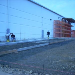 People walking beside a large white industrial building on a gravel lot.