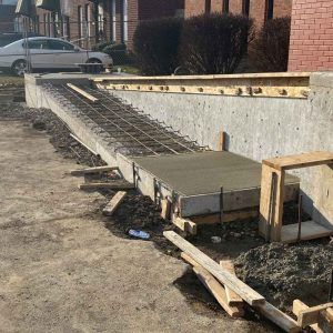 Damaged concrete ramp with exposed rebar and debris beside a brick building and parked car