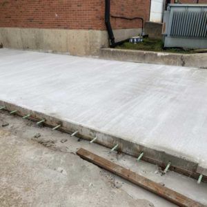 Freshly poured concrete slab beside a brick wall, with forms and a drain grate at the edge.