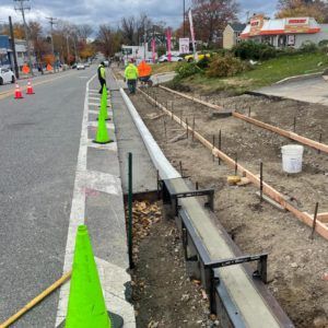 Roadside construction with orange cones, workers in safety vests, and an open curb trench with drainage pipes