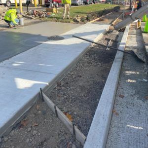 Workers pouring concrete for a sidewalk beside a curb and gravel trench