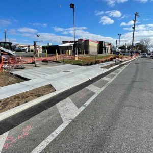 New concrete sidewalk ramp beside a street, with orange construction fencing and utility poles under a blue sky