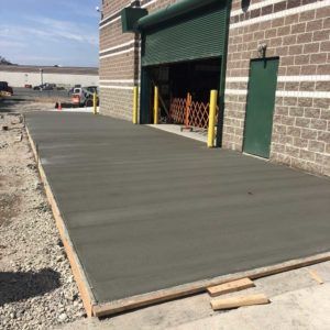 Freshly poured concrete ramp beside a brick building with a green roll-up door and gravel lot