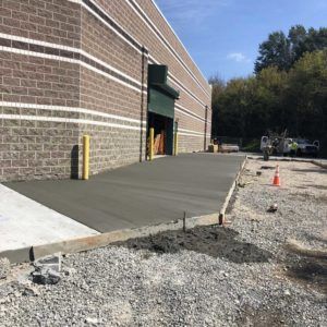 New concrete sidewalk beside a brick building, with an orange cone and gravel work area.
