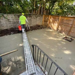 Worker pouring concrete in a backyard, standing on a long metal chute beside a fresh slab.