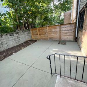 Concrete patio with a wooden privacy fence and trees in the background, seen from a doorway with a black railing.