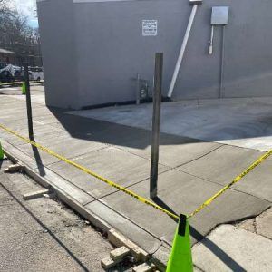 Caution-taped, uneven sidewalk ramp beside a gray building, with a green cone in the foreground