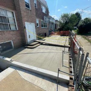 Construction site with freshly poured concrete patio beside a brick building, framed by orange safety fencing