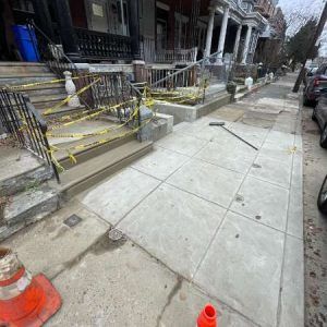 Sidewalk under construction with yellow caution tape, concrete panels, and orange traffic cones beside townhouses