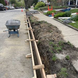 Sidewalk trench under construction with wooden bracing and a wheelbarrow on the pavement