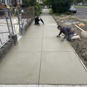 Workers smoothing fresh concrete on a sidewalk beside a chain-link fence and dirt strip
