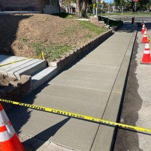 New concrete sidewalk under construction, bordered by caution tape and orange cones beside a grassy slope.