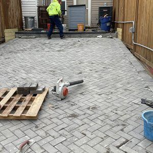 Worker paving a driveway with gray interlocking bricks, tools and pallets in the foreground.