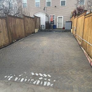 Empty brick-paved driveway between wooden fences leading to a house with two trash bins at the back.
