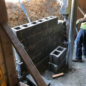 Construction worker building a cinder block wall in a trench beside an excavated dirt slope.
