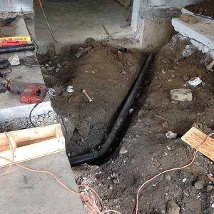 Indoor floor under renovation with exposed dirt trench and black pipe, surrounded by construction tools and debris