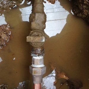 Metal pipe fitting standing in muddy brown water, with a leak or repair joint visible near the center