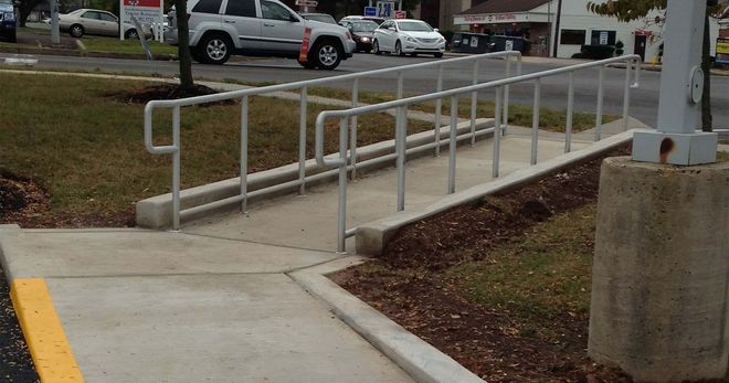 Concrete ramp with metal handrails leading up a sidewalk beside a parking lot.