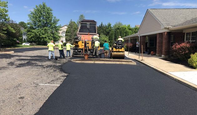 Road paving crew laying fresh black asphalt beside a building, with a paver and rollers on a sunny day