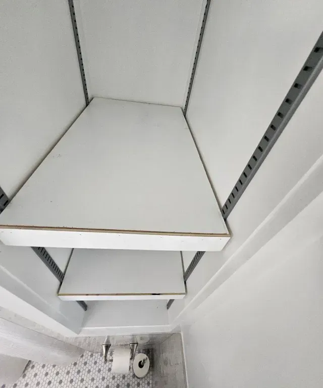 Empty white retail shelves with metal rails and a few items on the bottom shelf, viewed from above