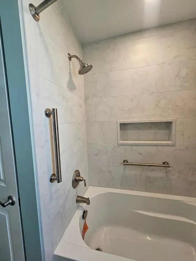 Walk-in shower with white tile, chrome grab bars, built-in shelf, and a tub edge visible at the bottom.