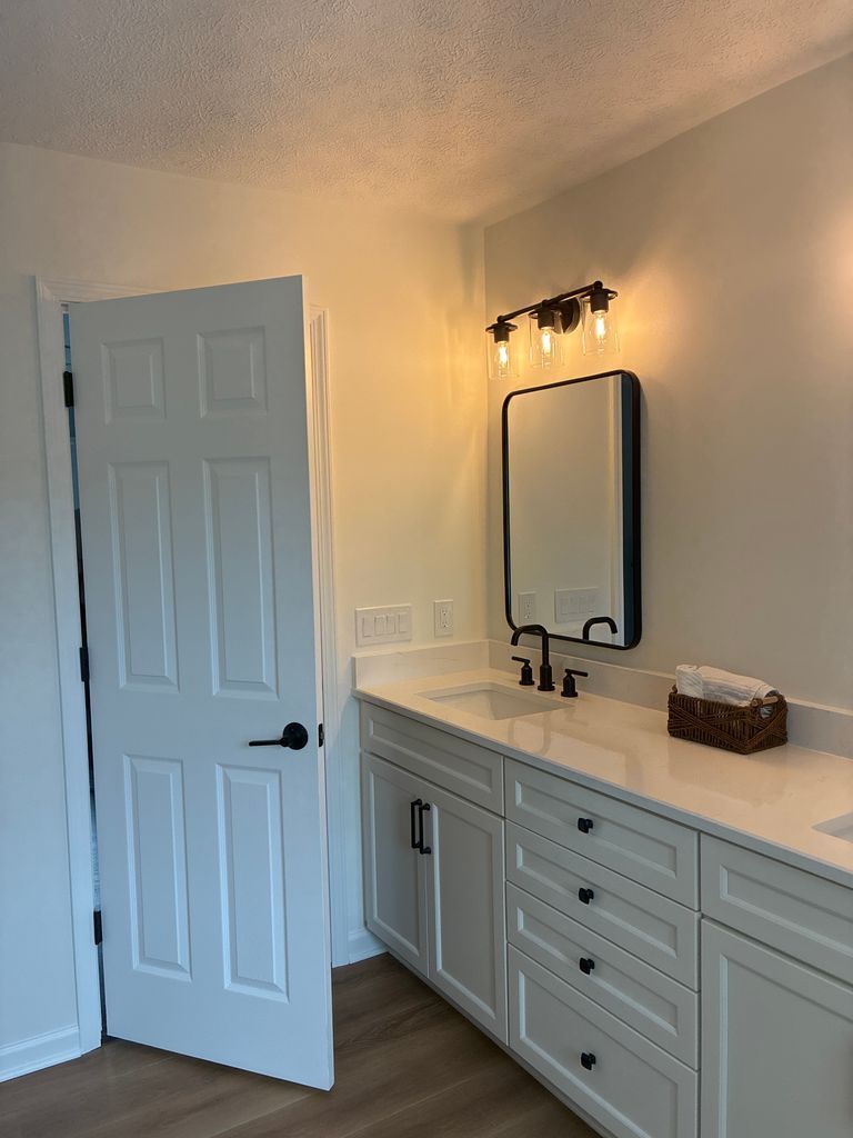 Bright bathroom vanity with mirror, black faucet, wall light, and white door ajar.