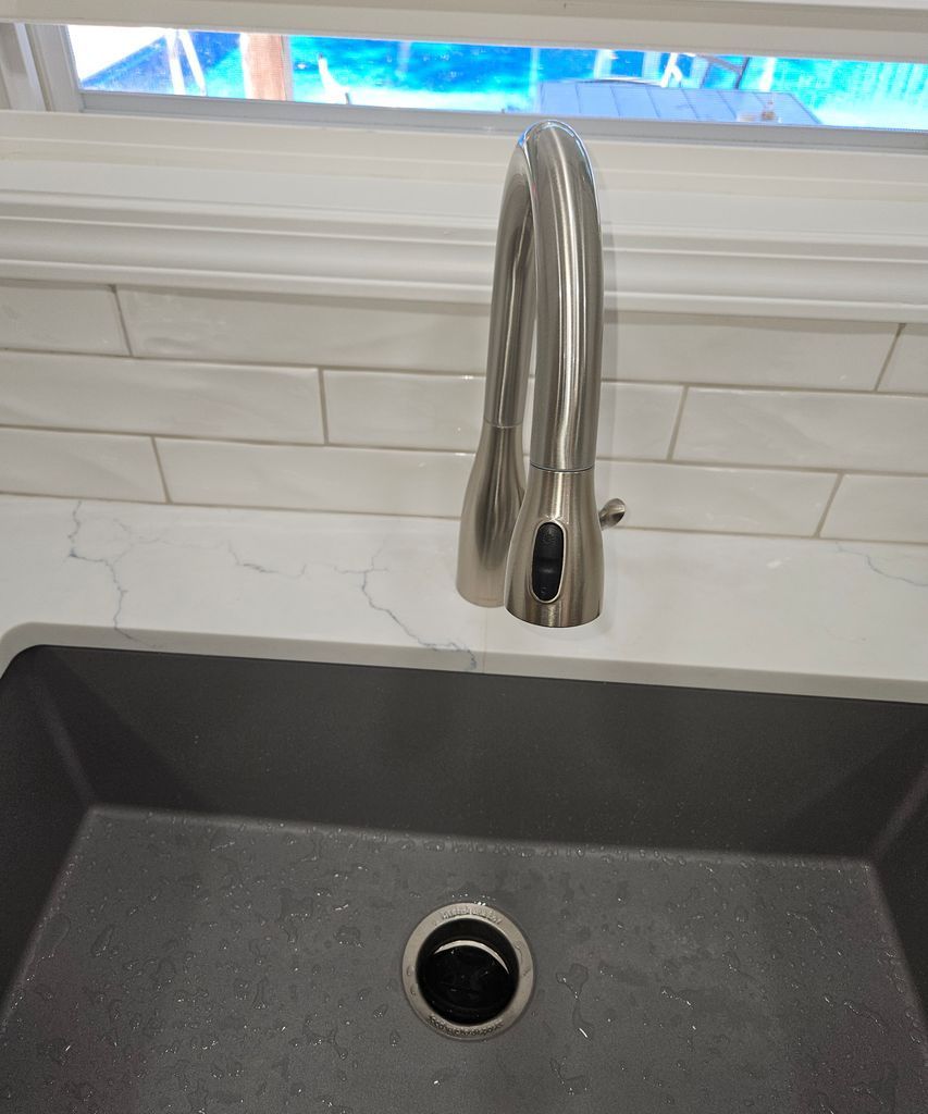 Stainless steel kitchen faucet over a dark sink with white tile backsplash and window above
