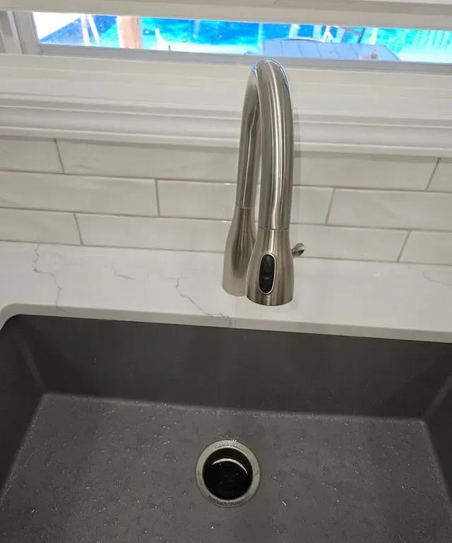 Stainless steel kitchen faucet over a black sink with a tiled backsplash and window above