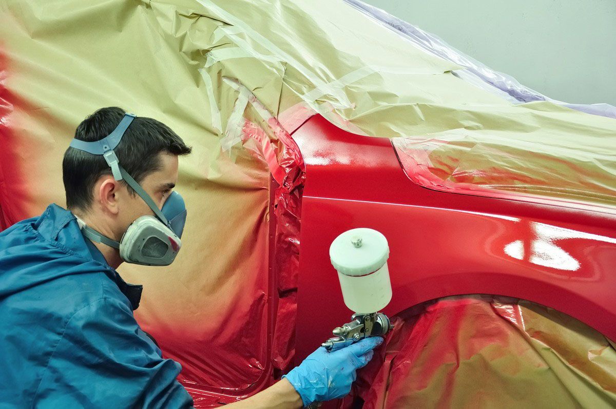 Man in protective gear spray-painting a red car in a workshop.