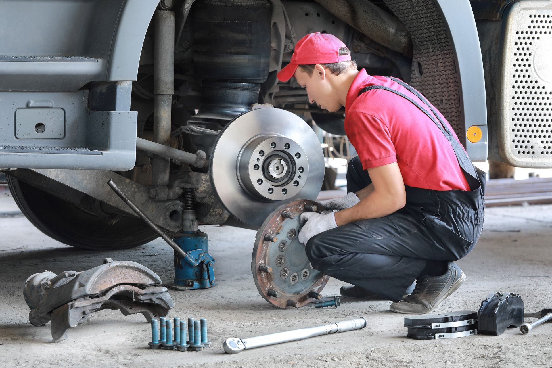 Mechanic working on truck brakes, wearing red shirt and cap. Shop setting, tools and parts visible.