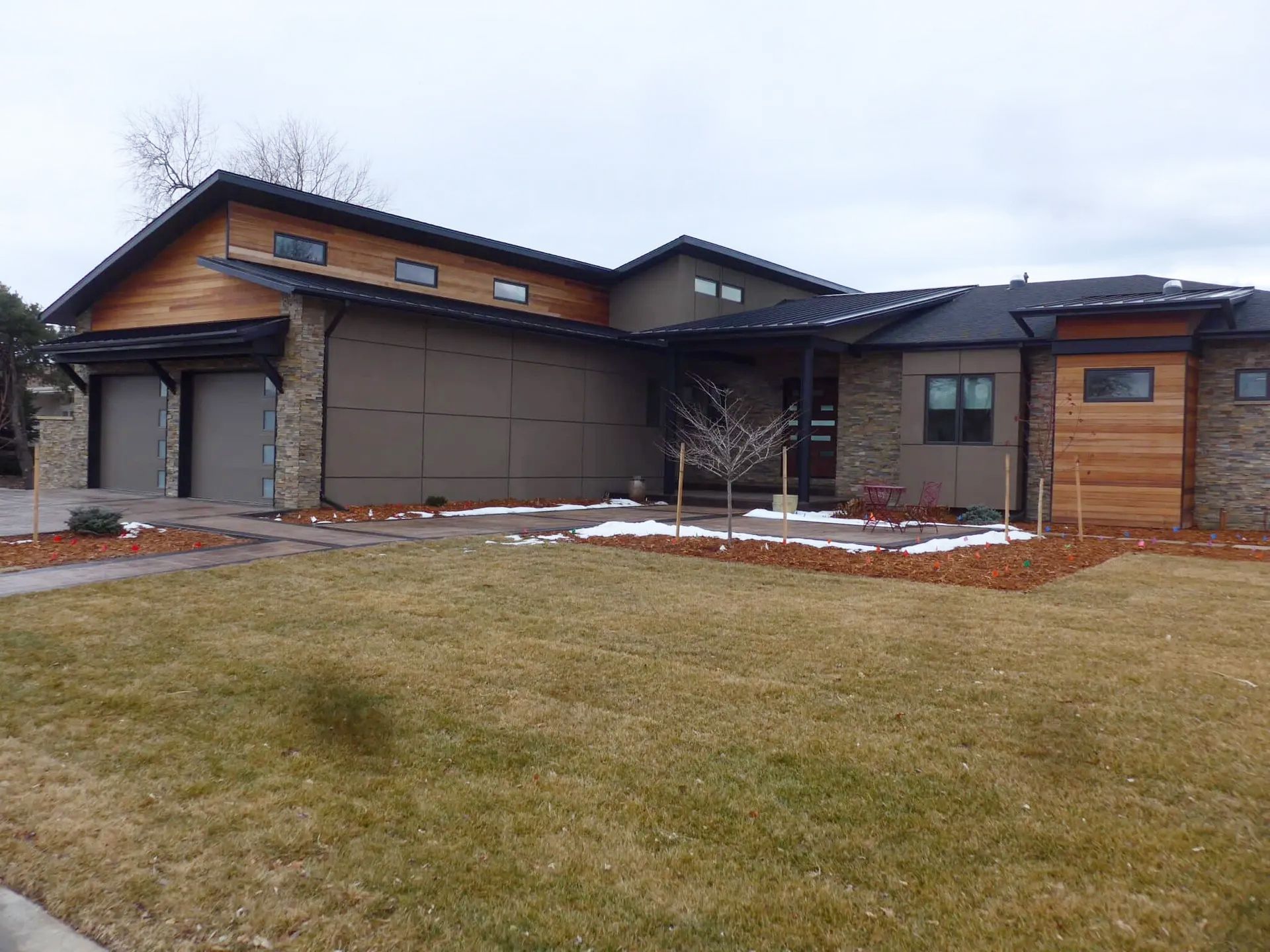 A modern house with a mix of stone, dark metal, and wood siding, featuring a two-car garage and a landscaped yard.