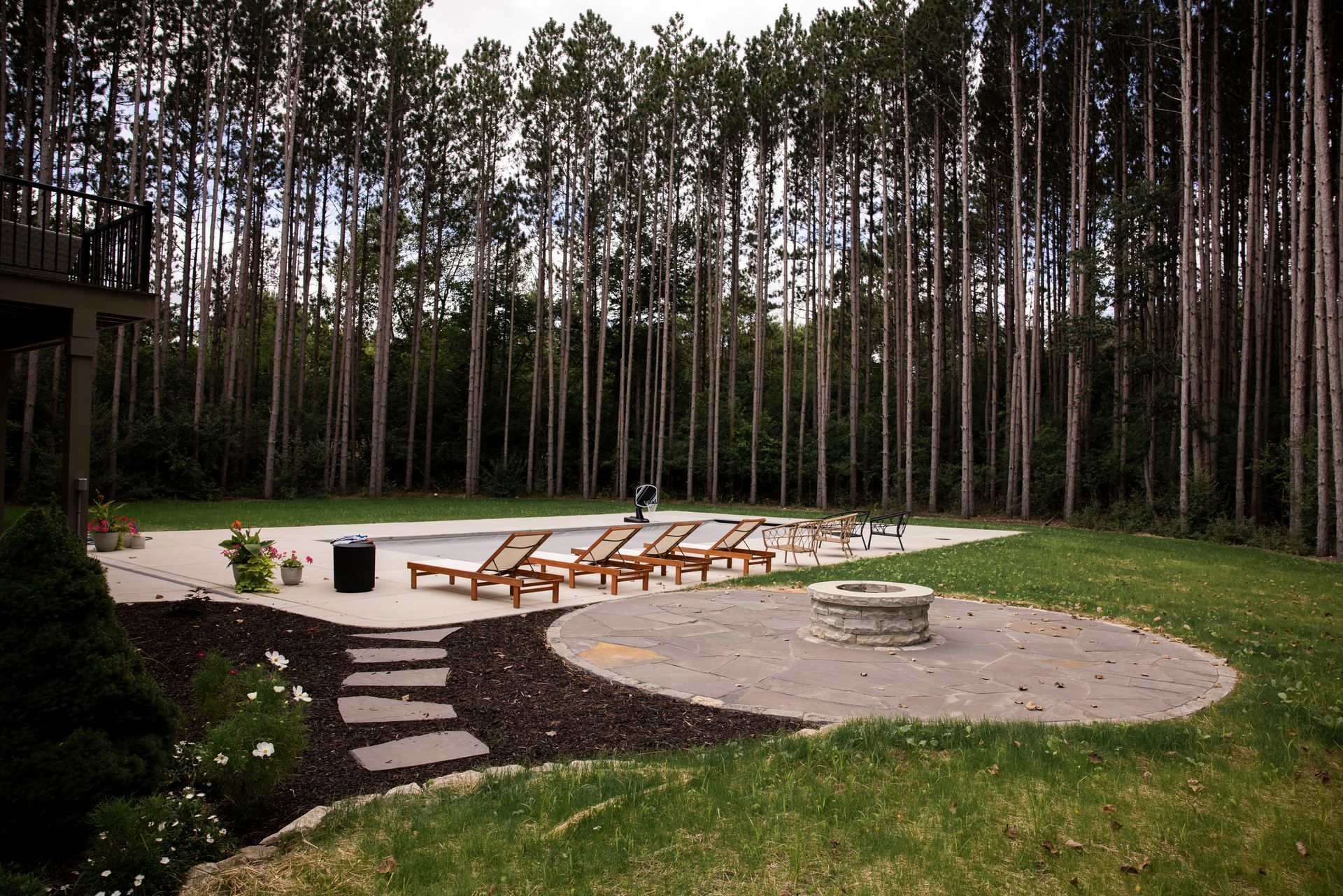 Backyard with a swimming pool, lounge chairs, and a circular stone fire pit, bordered by a dense forest of tall pines.