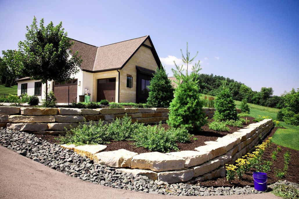 A stone house sits behind a tiered garden wall with evergreen trees, shrubs, and yellow flowers under a clear blue sky.