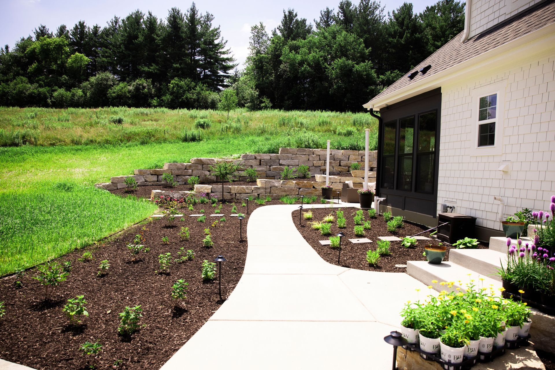 A white-walled house with a path leading through a mulched garden towards a hillside with terraced planting beds.