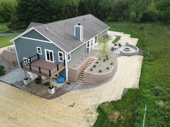 An aerial view of a gray house with a wooden deck, stone retaining wall, and a circular fire pit area in the backyard.