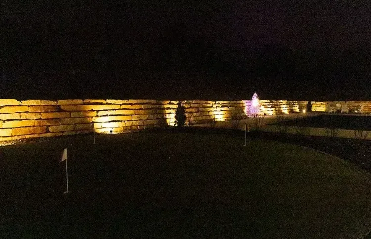 A putting green at night, illuminated by landscape lights highlighting a stacked stone retaining wall.